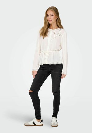 Weißes Langarmblusenshirt mit Spitzendetails und Knopfverschluss, kombiniert mit schwarzen Jeans mit Rissen und weißen Sneakers mit schwarzen Akzenten.