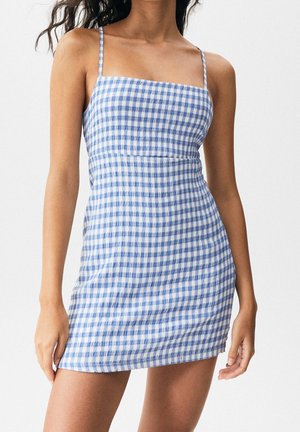 Kvinde iført en tætsiddende, blå- og hvide ternet minikjole med tynde skulderstropper foran en ensfarvet baggrund.