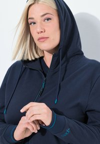 Felpa con zip di colore blu navy, dotata di cappuccio, con accenti e lacci turchesi a contrasto, realizzata in un morbido tessuto strutturato.