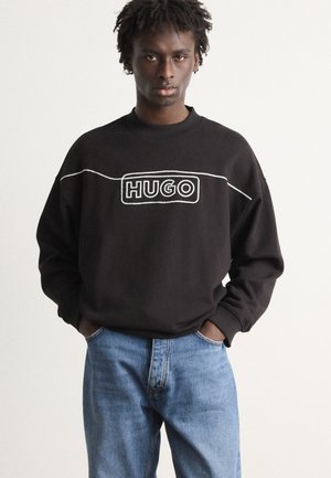 Juodas džemperis su baltu "HUGO" logotipu, apvaliu kaklu ir ilgomis rankovėmis, derinamas su šviesiai mėlynomis džinsinėmis kelnėmis.