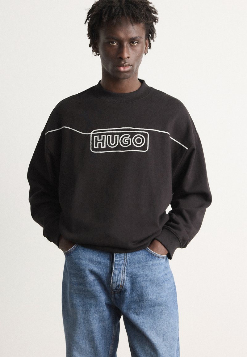 Fekete pulóver fehér "HUGO" logóval, kerek nyakkal és hosszú ujjakkal, világoskék farmernadrággal párosítva.