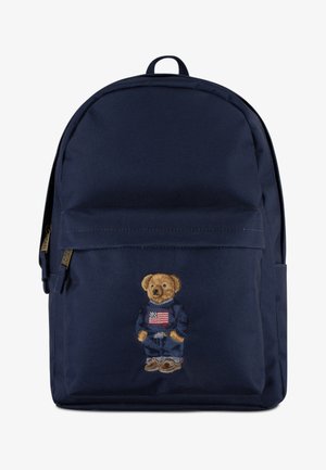 Zaino blu navy con un orsetto ricamato che indossa un maglione blu navy con una bandiera americana, in tessuto testurizzato e con una tasca frontale zip.