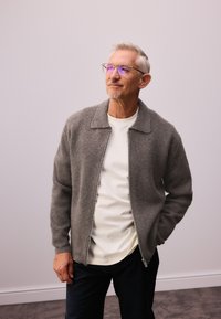 Grijze wollen ritssluiting jas met een kraaghals, gedragen over een crèmekleurig T-shirt en donkere broek. Geplaatst tegen een lichte achtergrond.