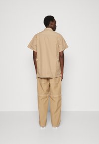 Tan short-sleeve shirt with a straight cut and button details, paired with loose tan pants featuring visible stitching and side pockets.