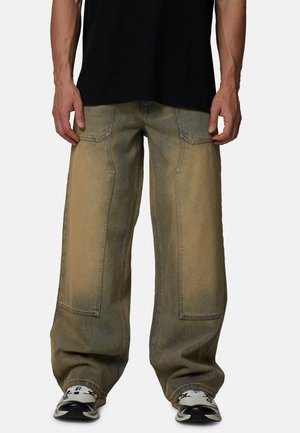 Losse spijkerbroek in een vervaagde lichtbruine kleur, voorzien van grote zakken en naden als detail. Draagt zwarte sneakers en een zwart shirt.