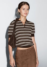 Brunstribet kortærmet polo-shirt med krave, parret med en brun ruskindsnederdel. Modellen bærer en matchende brun skuldertaske.
