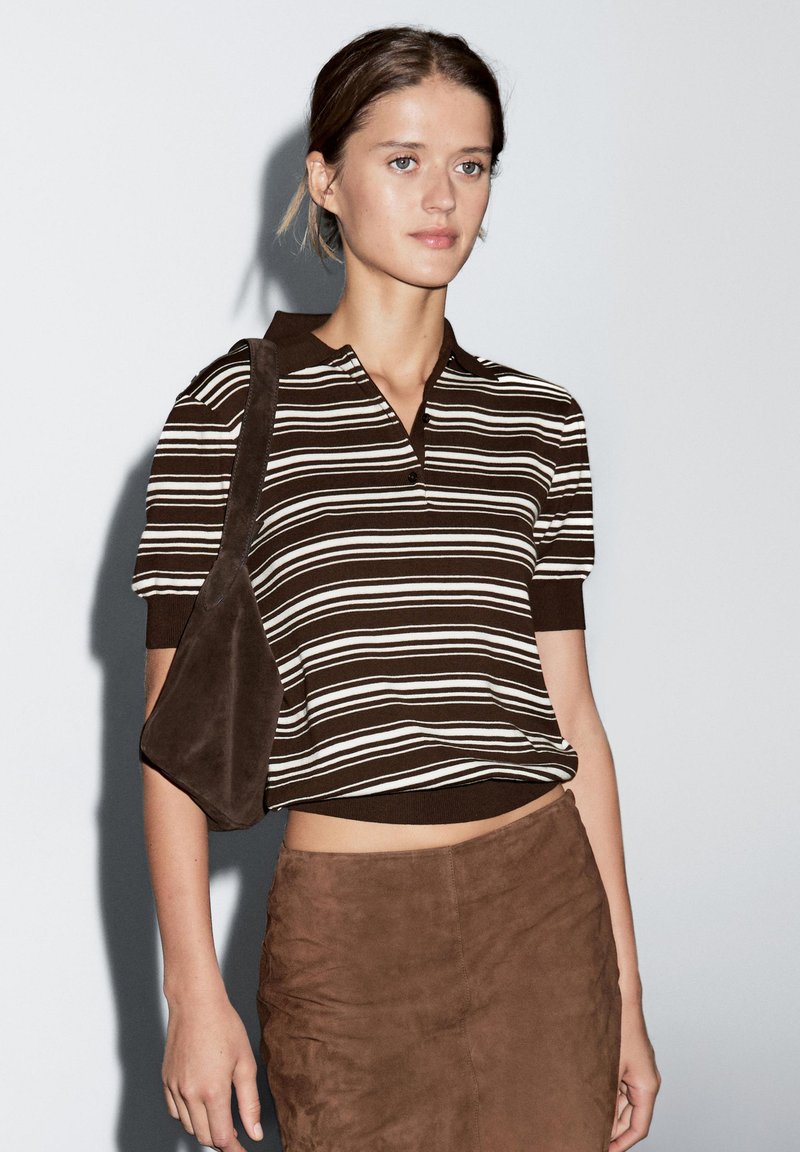 Brunstribet kortærmet polo-shirt med krave, parret med en brun ruskindsnederdel. Modellen bærer en matchende brun skuldertaske.