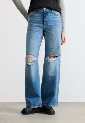 Άτομο που φοράει ανοιχτό μπλε, φαρδιά τζιν με σκισίματα στα γόνατα, μαύρα χοντρά παπούτσια, γκρι μακριά μανίκια και μαύρο τοπ.