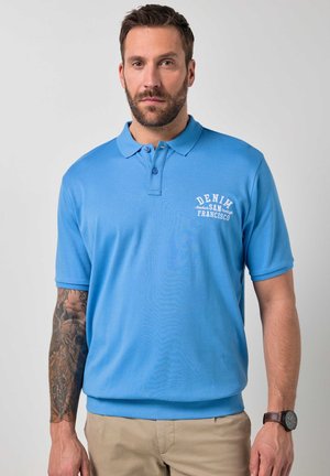 Mann mit Bart in blauem Poloshirt mit dem Text „DENIM SAN FRANCISCO“, Tattoo-Ärmel am linken Arm, beigefarbene Hose und Armbanduhr, stehend vor grauem Hintergrund.