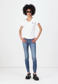 Balta V formos kaklo marškinėliai su maža juoda logo, derinami su šviesiai mėlynomis aptemptomis džinsų kelnėmis ir juodais platforminiais batais. Modelis su garbanotais plaukais ir akiniais nuo saulės.