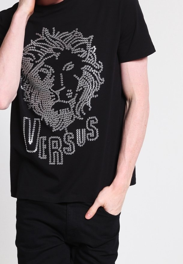 Personne portant un T-shirt noir avec une tête de lion argentée cloutée et le mot "VERSUS" sur le devant, main dans la poche.