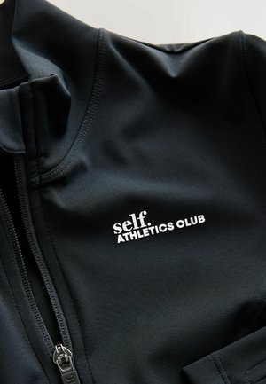 Close-up van een zwart sportjack met een rits en de tekst "self. ATHLETICS CLUB" prominent weergegeven in het wit.