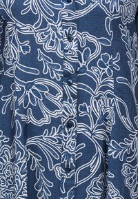 Μπλε ύφασμα με λευκά λουλουδένια και φυλλώδη σχέδια που περιβάλλουν μια κατακόρυφη σειρά από σκούρα κουμπιά στο κέντρο.