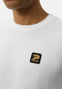 Weißes Baumwoll-T-Shirt mit Rundhalsausschnitt und einem schwarzen Quadrat-Patch mit einem goldenen Emblem und dem Text "PATRICK." Weicher Stoff, einfaches Design.