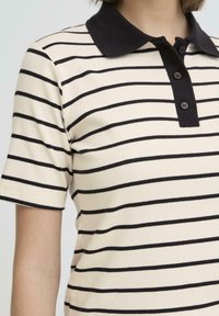 Personne portant un polo crème à manches courtes avec des rayures horizontales noires et un col noir uni à trois boutons.