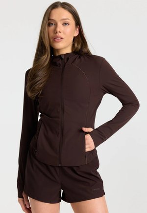 Jonge vrouw met lang bruin haar die een donkerbruine jas met rits en bijpassende korte broek draagt tegen een effen lichte achtergrond.