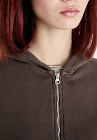 Hnedá mikina na zips s kontrastným strieborným zipsom. Mäkká látka s hladkou textúrou, má kapucňu a jednoduchosť dizajnu.