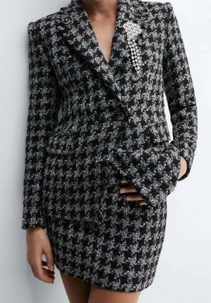 Femme portant un blazer et une jupe à motif pied-de-poule noir et blanc, avec une broche en perle épinglée sur le revers du blazer.