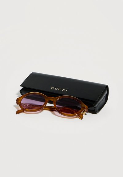 Lunettes de soleil ovales marron avec des verres violets reposant devant un étui Gucci noir avec des lettres dorées sur une surface blanche.