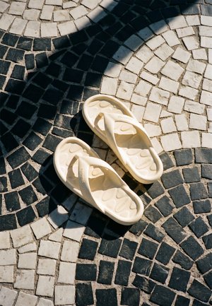 Tongs couleur crème reposant sur un pavement circulaire en carreaux noirs et blancs avec des motifs de lumière et d’ombre.