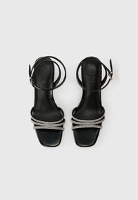 Melnas augstpapēžu sandales ar kvadrātu purngalu, kurām ir trīs rhinestones rotātas jostas un plānas potītes jostas. Gluda ādas tekstūra.