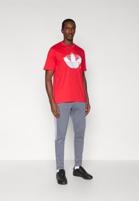 Κόκκινο βαμβακερό T-shirt με μεγάλο λευκό γραφικό λογότυπο, σε συνδυασμό με γκρι φόρμα με λευκές πλευρικές ρίγες και μαύρα αθλητικά παπούτσια.