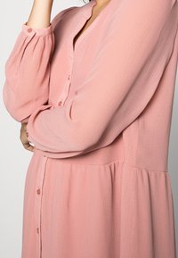 Langmouw roze jurk van lichtgewicht stof met een gestructureerd oppervlak. Bevat knoopdetails en een ontspannen pasvorm met een gathered taille.