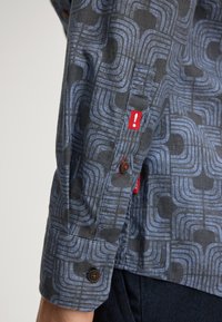 Langzaam shirt met een geometrisch blauw en grijs patroon, met rode accenten en bruine knopen op de manchetten. Van katoen.