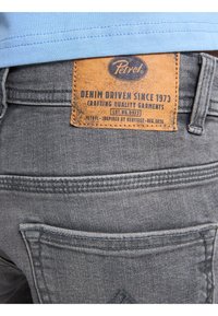 Jeans de mezclilla gris con una etiqueta de piel que muestra el logo "Petrol", texto y acentos naranjas. Presenta costuras en los bolsillos traseros y un acabado texturizado.