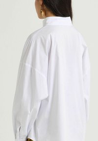 Camicia bianca in cotone, taglio ampio, con colletto alto, spalle scese e spacco laterale sull'orlo. Tessuto liscio, design minimalista.