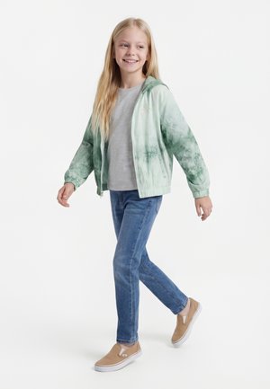 Dziewczyna o długich blond włosach idzie, ubrana w zieloną bluzę na suwak z motywem tie-dye, szarą koszulkę, niebieskie dżinsy i beżowe buty wsuwane na białym tle.