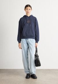 Sweatshirt com capuz azul marinho com um logótipo, jeans azul claro, mala preta e sapatos pretos. Conjunto casual com um corte descontraído e design minimalista.