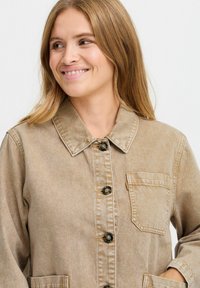 Beige denimjakke med krave, knaplukning, brystlomme og synlige syningdetaljer. Blødt materiale med afslappet pasform.