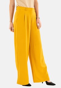 Pantaloni a gamba larga in tessuto giallo vivace, con pieghe frontali e tasche laterali. La texture è liscia, abbinata a tacchi a punta neri.