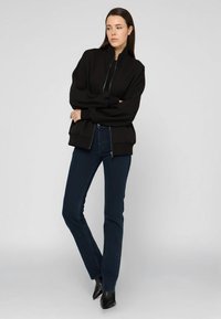 Černá bomber bunda na zip s vysokým límcem, oversized rukávy, v kombinaci s tmavě modrými džíny rovného střihu a černými botami na podpatku s špičatou špičkou.