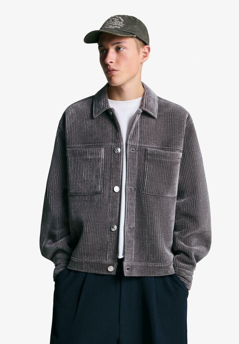 Szara kurtka sztruksowa z kołnierzem, zapinana na guziki z przodu, z dwoma kieszeniami na piersi i długimi rękawami. Noszona na białej koszuli i zestawiona z ciemnymi spodniami.