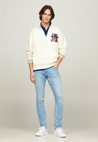 Helle beige Sweatshirt mit halbem Reißverschluss und gesticktem Logo, gestreiften Bündchen und navyblauem Poloshirt darunter. Kombiniert mit hellblauen Jeans und weißen Sneakern.