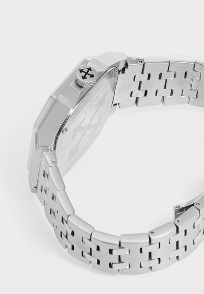 Montre en métal argenté avec bracelet à maillons et dos gravé comportant une couronne avec un logo à quatre flèches sur le côté.