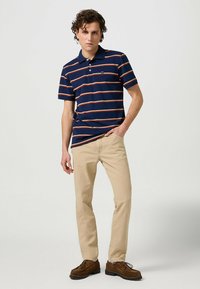Granatowa koszulka polo w pomarańczowe poziome paski, z krótkimi rękawami i trzema guzikami, w połączeniu z beżowymi spodniami chinos i brązowymi butami.