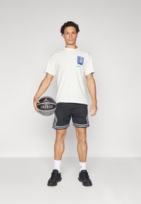Light beige T-shirt with blue graphic and Nike logo, black mesh shorts with white stripes, holding a black basketball with red details.
