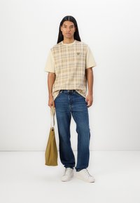 Kortermet rutete skjorte i beige og kremfarge med rund hals, kombinert med blå jeans og hvite sneakers. Holder en beige skulderveske.