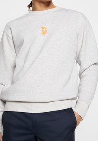 Person som bär en ljusgrå sweatshirt med en liten orange grafik på bröstet och mörkblå byxor, handen i fickan, mot en vit bakgrund.