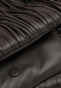 Sac en cuir marron avec détails à franges, texture lisse et fermeture visible à bouton-pression rond. Gros plan mettant en valeur le contraste des matériaux.