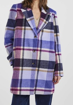 Femme portant un manteau à carreaux violet, bleu et beige avec des boutons, debout devant un fond uni.