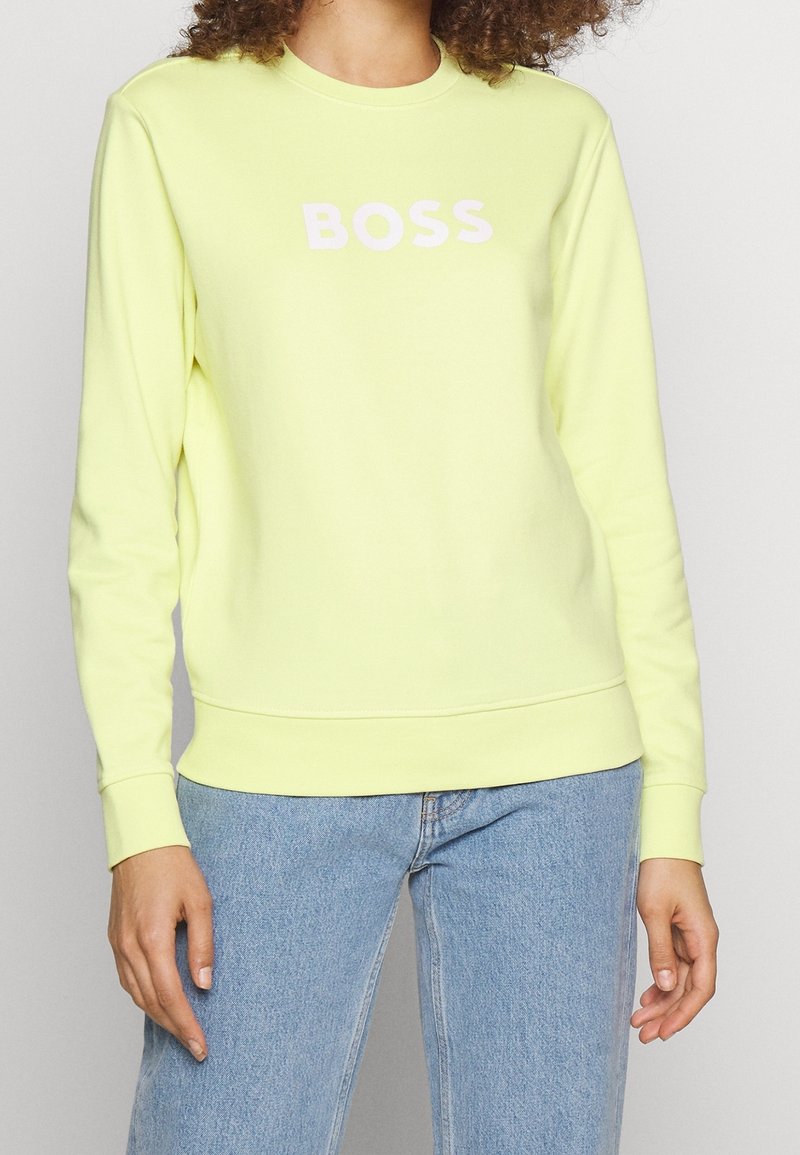 Felpa gialla con scollo rotondo, maniche lunghe e polsini a costine. Presenta un grande logo "BOSS" in rosa chiaro al centro.