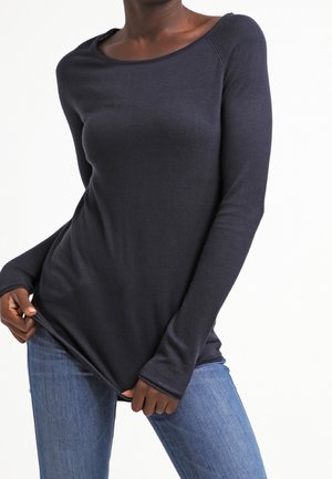 Frau trägt ein eng anliegendes langärmeliges dunkelblaues Oberteil und blaue Jeans, zieht den Saum des Oberteils leicht nach außen.