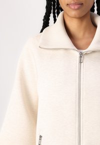 Lys beige lynlåsjakke lavet af blødt stof, med en ribbet krave og to sidelommer med lynlås. Glat tekstur og moderne design.