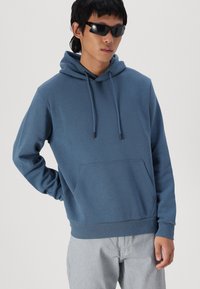 Felpa blu in cotone con tasca frontale, coulisse regolabile e polsini a coste; indossata con pantaloni grigi a trama. Il modello indossa occhiali da sole.
