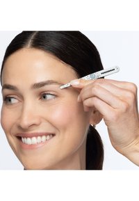 Donna sorridente mentre si sistema le sopracciglia con pinzette bianche Tweezerman vicino all'occhio destro, su sfondo bianco uniforme.