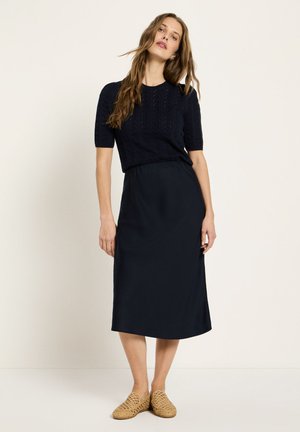 Woman with long hair wearing a black textured short-sleeve top, black midi skirt, and woven beige slip-on shoes, standing against a plain background.